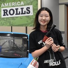A photo of a high-school aged young woman wearing a TechForce t-shirt and holding a Snap-on Impact Driver next to TechForce's NASCAR Clip Car Challenge, part of our Mobile STEM Career Center.